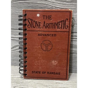 May include: A spiral-bound notebook with a brown fabric cover featuring the black text THE STONE ARITHMETIC, ADVANCED, and STATE OF KANSAS. A circular geometric logo is centered on the front cover, and a small white dot appears near the top.