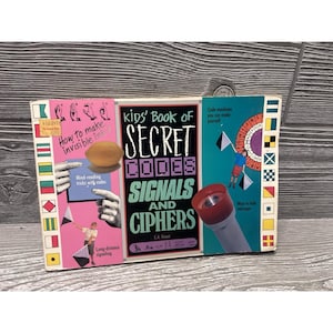 May include: A vintage children's book titled "Kids' Book of Secret Codes, Signals and Ciphers" with colorful illustrations. The book cover features text and images related to codes, signals, and invisible ink. The book is displayed on a wooden surface.