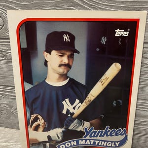 May include: A Topps baseball card featuring Don Mattingly of the New York Yankees. The card has a red border and displays the Yankees logo. Mattingly is wearing a navy blue jersey and a black cap, holding a baseball bat and wearing a glove.