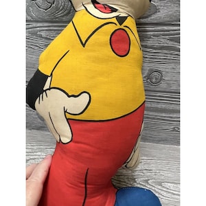 May include: Vintage plush toy of Mickey Mouse. The toy is made of fabric and has a yellow shirt, red pants, and black shoes. The toy is in good condition and has a classic look.