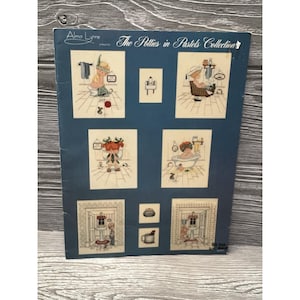 May include: A blue poster featuring cross-stitch patterns from the "Alma Lynne presents The Potties in Pastels Collection." The patterns depict bathroom scenes with figures in pastel colors. The poster is on a wooden surface.