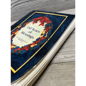 May include: A blue book with a gold border and a wreath of red berries, yellow pears, and blue grapes. The book title is "150 Years of Blessings" and the text "First United Methodist Church, Athens, Texas" is below the wreath.