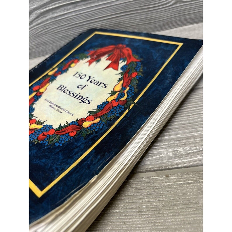 May include: A blue book with a gold border and a wreath of red berries, yellow pears, and blue grapes. The book title is "150 Years of Blessings" and the text "First United Methodist Church, Athens, Texas" is below the wreath.