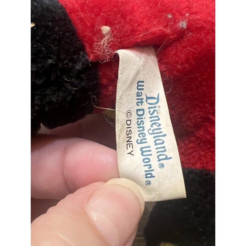May include: Close-up of a vintage plush toy with red and black fur. A white tag with the words "Disneyland Walt Disney World" and the Disney logo is visible. The toy's fur appears worn.