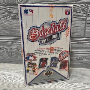 Puede incluir: Una caja sellada de cartas de béisbol de la edición de 1991. La caja es blanca con detalles en rojo y azul, con la palabra "Baseball" en una fuente estilizada. La caja incluye imágenes de jugadores de béisbol y el texto "The Collectors Choice."