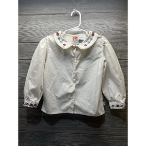 May include: Off-white long-sleeve button-down shirt for children. The collar and cuffs are adorned with embroidered floral patterns in red, blue, and green. The shirt has the text "McDonald's Kids" on the collar.