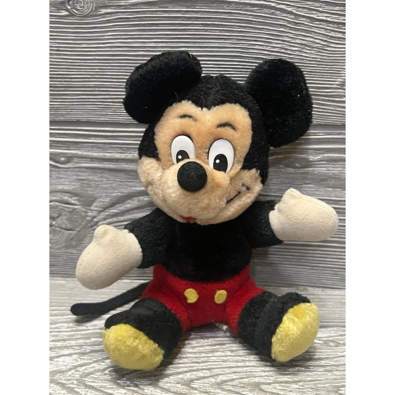 May include: A plush Mickey Mouse toy. It has black fur, large black ears, and a tan face. He wears red shorts with yellow buttons, white gloves, and yellow shoes. The toy is sitting with its arms outstretched.