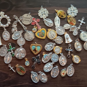 May include: A collection of silver and gold religious charms, including crosses, hearts, and medallions with images of saints. The charms are arranged on a wooden surface.