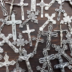 May include: A collection of silver-colored crucifix pendants, each with intricate details and varying designs. The crosses are displayed on a dark wooden surface, showcasing their religious theme and craftsmanship.