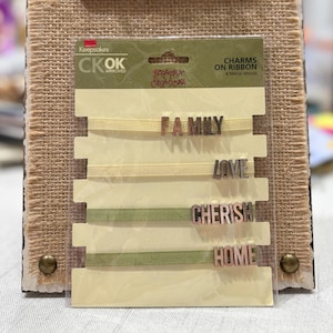 May include: A clipboard with a burlap-textured background and a metal clip. A package of ribbon charms with the words "FAMILY", "LOVE", "CHERISH", and "HOME" is attached to the clipboard. The package reads "CHARMS ON RIBBON 4 Metal Words".