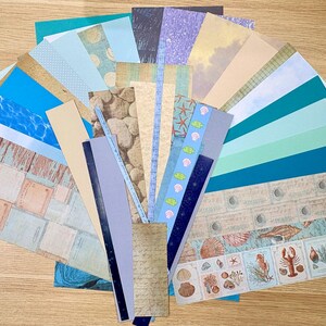May include: A collection of patterned paper sheets in various shades of blue, teal, and beige, arranged in a fan shape. Designs include ocean waves, seashells, and vintage-style graphics. Ideal for scrapbooking and crafting projects.