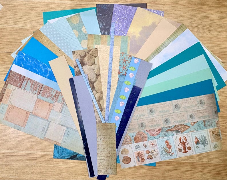 May include: A collection of patterned paper sheets in various shades of blue, teal, and beige, arranged in a fan shape. Designs include ocean waves, seashells, and vintage-style graphics. Ideal for scrapbooking and crafting projects.