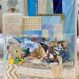 May include: A collection of scrapbook paper and embellishments in a clear plastic bag. The kit includes patterned paper, lace trim, paper flowers, a butterfly, and other decorative elements in shades of blue, beige, and white.