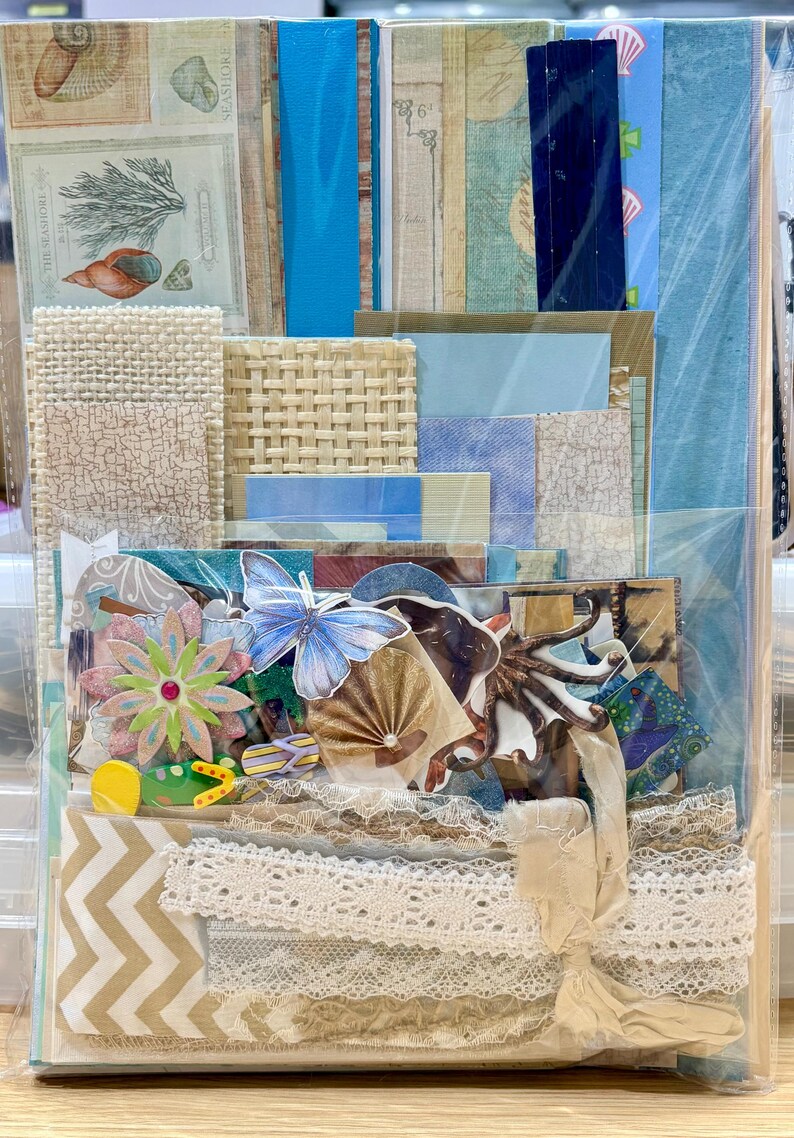 May include: A collection of scrapbook paper and embellishments in a clear plastic bag. The kit includes patterned paper, lace trim, paper flowers, a butterfly, and other decorative elements in shades of blue, beige, and white.