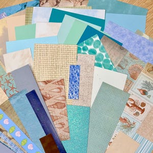 May include: A collection of various paper sheets in shades of blue, beige, and green. Some sheets feature seashell and starfish patterns, while others have textured or patterned designs. The assortment includes solid colors and patterned papers.