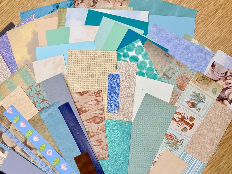 May include: A collection of various paper sheets in shades of blue, beige, and green. Some sheets feature seashell and starfish patterns, while others have textured or patterned designs. The assortment includes solid colors and patterned papers.