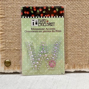 May include: A package of rhinestone accents in the shape of the letter "W". The accents are clear and pink rhinestones. The package is on a light green background with white polka dots. The top of the package has a black background with cherries.
