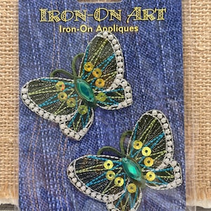 May include: Two iron-on butterfly appliqués. The butterflies have black, teal, and yellow details with a green gem in the center. The edges are outlined with white beads. The package reads "IRON-ON ART" and "Iron-On Appliques".