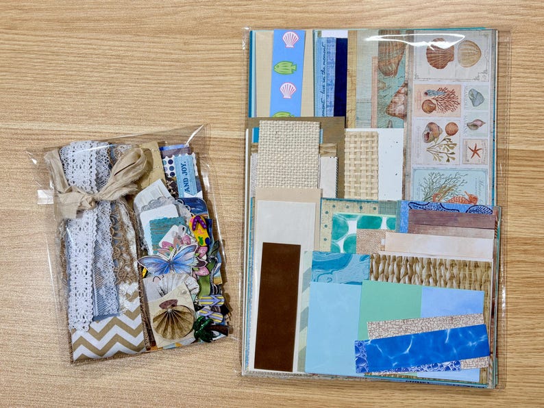 May include: Two packages of craft supplies. One is a clear bag tied with twine, containing lace, paper, and embellishments. The other is a clear bag with various patterned papers, including seashell and wave designs, and the text "AND JOY."