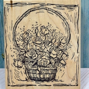 May include: A wooden stamp featuring a detailed illustration of a basket overflowing with roses. The design is rendered in black ink, showcasing the texture of the roses and the woven basket. The stamp is set against a light wood background.