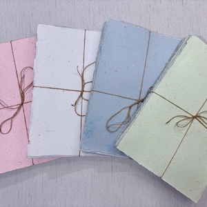 May include: Four stacks of handmade paper in pastel colors: pink, white, light blue, and pale green. Each stack is tied with twine in a crisscross pattern, creating a rustic and natural aesthetic. The paper has a textured, organic appearance.