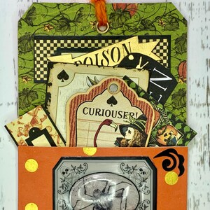 May include: A Halloween-themed tag with a pocket that opens to reveal a spider. The tag is decorated with a variety of vintage-style paper scraps, including a card that says "Curiouser!" and a tag that says "Poison".