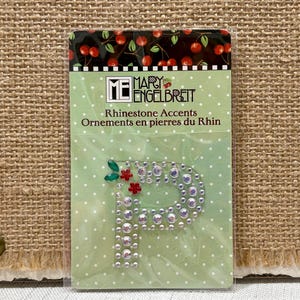 May include: A package of rhinestone accents featuring the letter "P". The letter is embellished with clear rhinestones and small red and green floral accents. The package has a green background with white polka dots and the text "Mary Engelbreit".