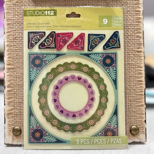 May include: A package of Studio 112 adhesive chipboard embellishments. The package contains nine pieces with floral and geometric designs in shades of green, pink, and blue. The package is displayed on a clipboard with a burlap texture.