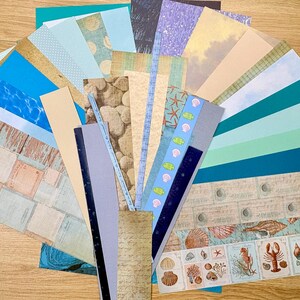 May include: A collection of patterned paper sheets in various colors and designs, including blue water, wood grain, and seashell motifs. The papers are arranged in a fan shape, suitable for scrapbooking or crafting projects.