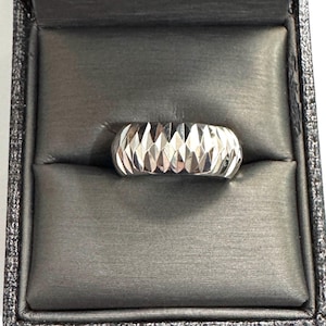 May include: A silver ring with a geometric, faceted design. The ring is displayed in a dark gray ring box with a textured interior. The ring's surface reflects light, creating a sparkling effect. The ring is a band style.