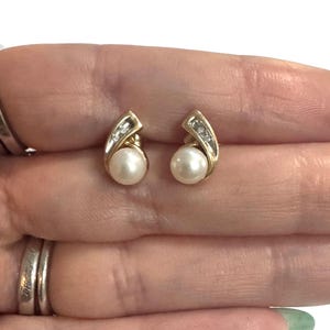 May include: A pair of gold stud earrings, each featuring a round white pearl set at the base of a curved gold accent piece. The curved gold section is embellished with three small, clear, sparkling stones arranged in a vertical line.