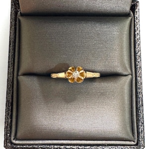 May include: A gold ring featuring a central flower-shaped setting with a small, clear, round-cut stone at its core. The band has a smooth, polished finish with decorative etched lines on the shoulders, presenting a refined and classic appearance.