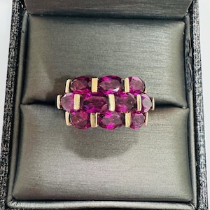 Könnte beinhalten: Ein goldener Ring mit drei Reihen oval geschliffener, tiefmagentafarbener Edelsteine. Die Edelsteine sind in einem goldenen Band gefasst, wodurch ein kühnes Design entsteht. Der Ring wird in einer schwarzen Schmuckschatulle präsentiert.