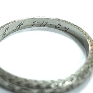 Puede incluir: Primer plano de un anillo de plata con detalles grabados. El anillo tiene la inscripción "A. 1-11-24" grabada en su superficie. La textura del anillo parece martillada o texturizada, lo que aumenta su atractivo vintage. El anillo está sobre un fondo blanco.