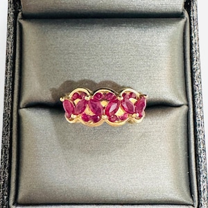 May include: A gold ring featuring a series of marquise-cut red rubies arranged in a cluster pattern. The band has a scalloped edge design that follows the shape of the gemstone settings, creating a continuous row of vibrant red stones across the top.