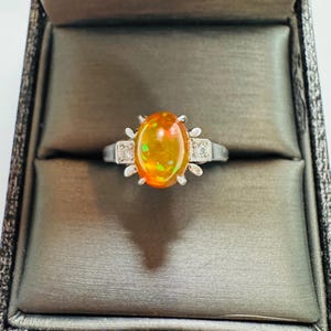 May include: A silver-toned ring featuring a central oval-shaped orange opal with iridescent green and yellow flashes. The setting includes small clear accent stones on either side of the opal, held in place by a prong setting on a metal band.