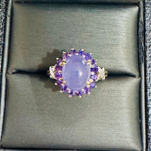 Puede incluir: Un anillo con una gran piedra ovalada de color lavanda, rodeada de piedras moradas más pequeñas. El engaste del anillo incluye detalles dorados y se presenta en una caja de joyería gris oscuro, resaltando su elegante diseño.