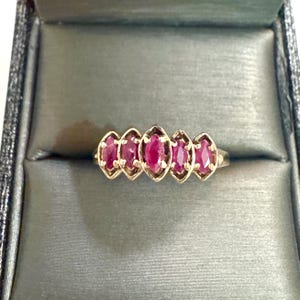 May include: A gold ring featuring five marquise-cut red gemstones arranged in a horizontal row. Each gemstone is held in a prong setting, with the center stone being slightly larger than the four flanking stones, creating a symmetrical design.