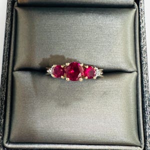 May include: A gold ring featuring a large round-cut vibrant red gemstone in the center, flanked by two smaller round-cut red gemstones. The setting includes tiny clear accent stones on each side of the band, creating a multi-stone design.