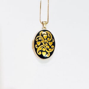 Puede incluir: Colgante de medallón ovalado dorado con un fondo de esmalte negro y un diseño de filigrana dorada. La filigrana presenta motivos de corazón y remolino. El medallón cuelga de una delicada cadena dorada, creando una pieza de joyería clásica y elegante.