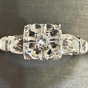 May include: A silver ring with a square-shaped center featuring a four-leaf clover design with a single diamond in the center.