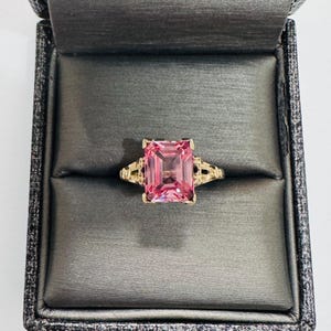 May include: A gold ring featuring a large, rectangular pink gemstone. The ring is presented in a dark grey jewellery box. The ring's band has decorative filigree details on each side of the gemstone.