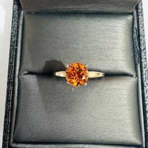 Puede incluir: Un anillo de oro con una gema naranja grande y redonda. El anillo está engastado en un engaste de cuatro puntas y se muestra en una caja de anillos gris.