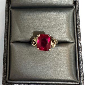 May include: A gold ring featuring a large, rectangular, faceted deep red gemstone held by four prongs. The band displays intricate, swirling metalwork on both sides of the central stone, creating a detailed and ornate appearance for the jewelry piece.