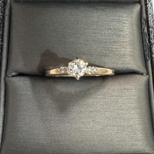 May include: A gold ring with a large, pear-shaped diamond center stone. The ring has smaller diamonds on either side of the center stone. The ring is set in a dark gray box.
