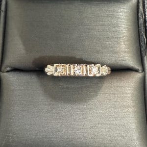 May include: A silver-toned metal ring featuring three small, clear, round-cut stones set in square-shaped bezels. The band has a decorative, fan-like engraving on both sides of the central stone setting, creating a symmetrical, detailed appearance.