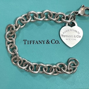 May include: A silver chain bracelet with a heart-shaped tag. The tag is engraved with "PLEASE RETURN TO TIFFANY & Co. NEW YORK". The bracelet is made of large, round links. The brand name "TIFFANY & Co." is also visible.