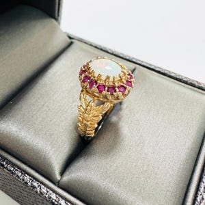 May include: A gold ring featuring a central oval cabochon opal surrounded by a halo of small round red rubies. The ring band is crafted with a textured, leaf-like design that extends from the base of the gemstone setting down the sides.