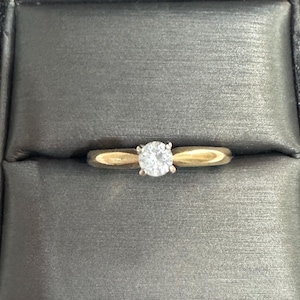 May include: A gold ring with a single, round, clear gemstone set in the center. The ring is set in a dark gray jewelry box. The band of the ring is a light gold color.