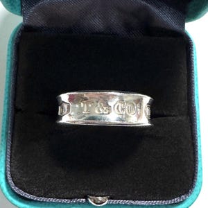May include: A silver ring with the text "T & CO" engraved on the band. The ring is displayed inside a teal blue jewelry box with a black interior.